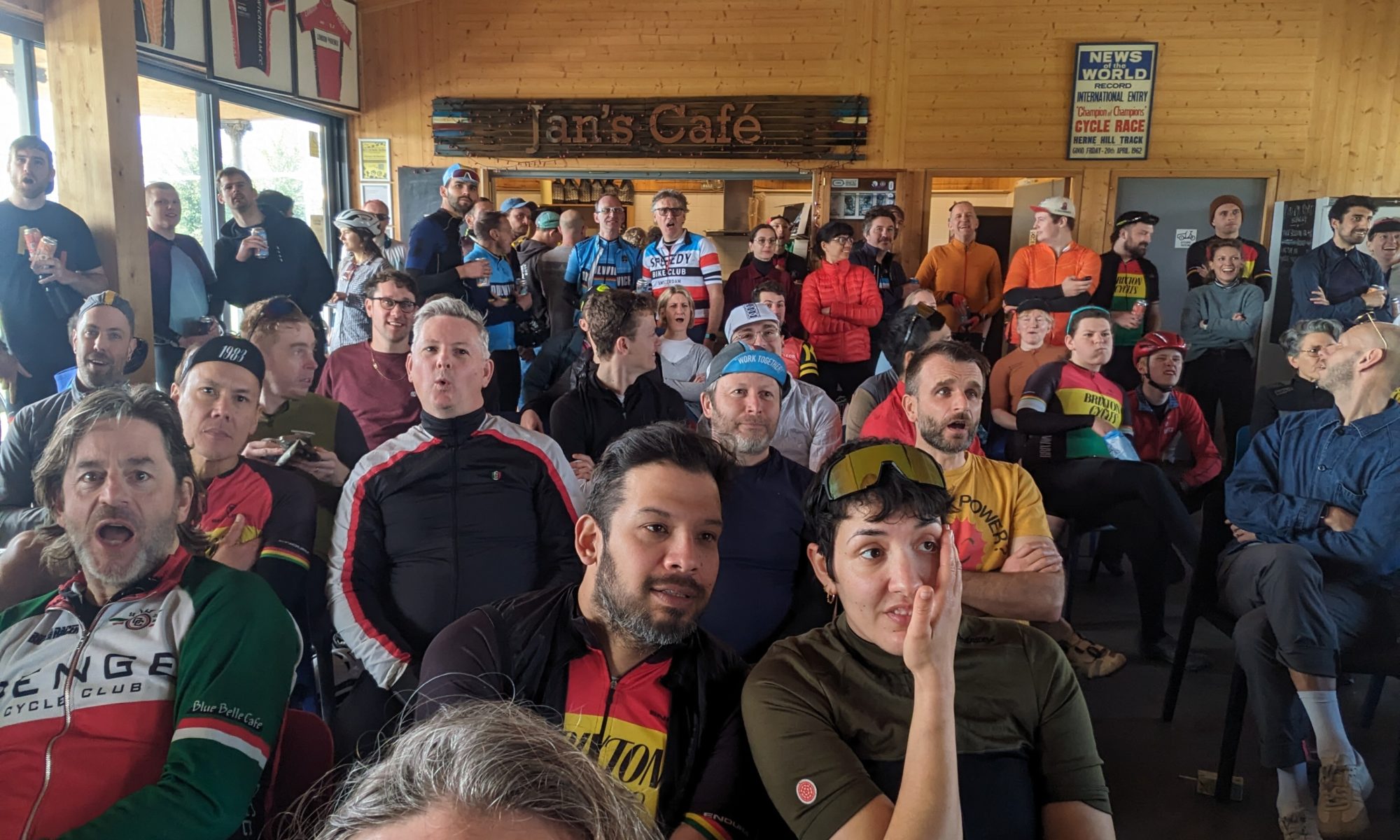 A crowd of cyclists are sitting in the HHV cafe reacting in shock to something happening during the men's Paris Roubaix race on TV.